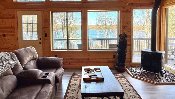 Interior de una cabaña de madera con sofá, mesa, chimenea y ventanas grandes con vistas a un lago.