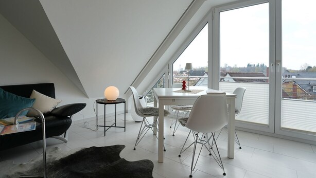 A modern loft bedroom with a sloped ceiling, large windows, a dining table, and a cozy seating area.