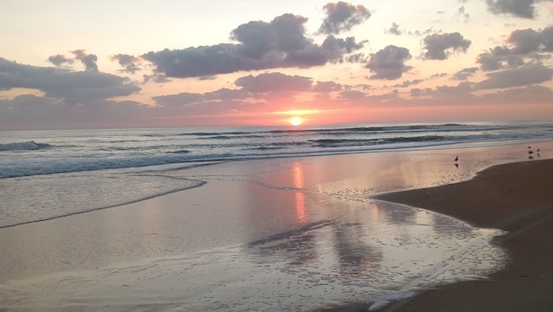 Una playa con arena mojada reflejando el atardecer, olas suaves lamiendo la orilla, y algunas personas caminando por la playa.