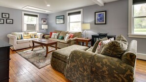 A living room with a white sofa, a beige sofa, and a camouflaged sofa. There are two windows, a lamp, and a rug. - Vermont Vacation Rental: Hot Tub, Near Ski Resorts (Mt Holly)