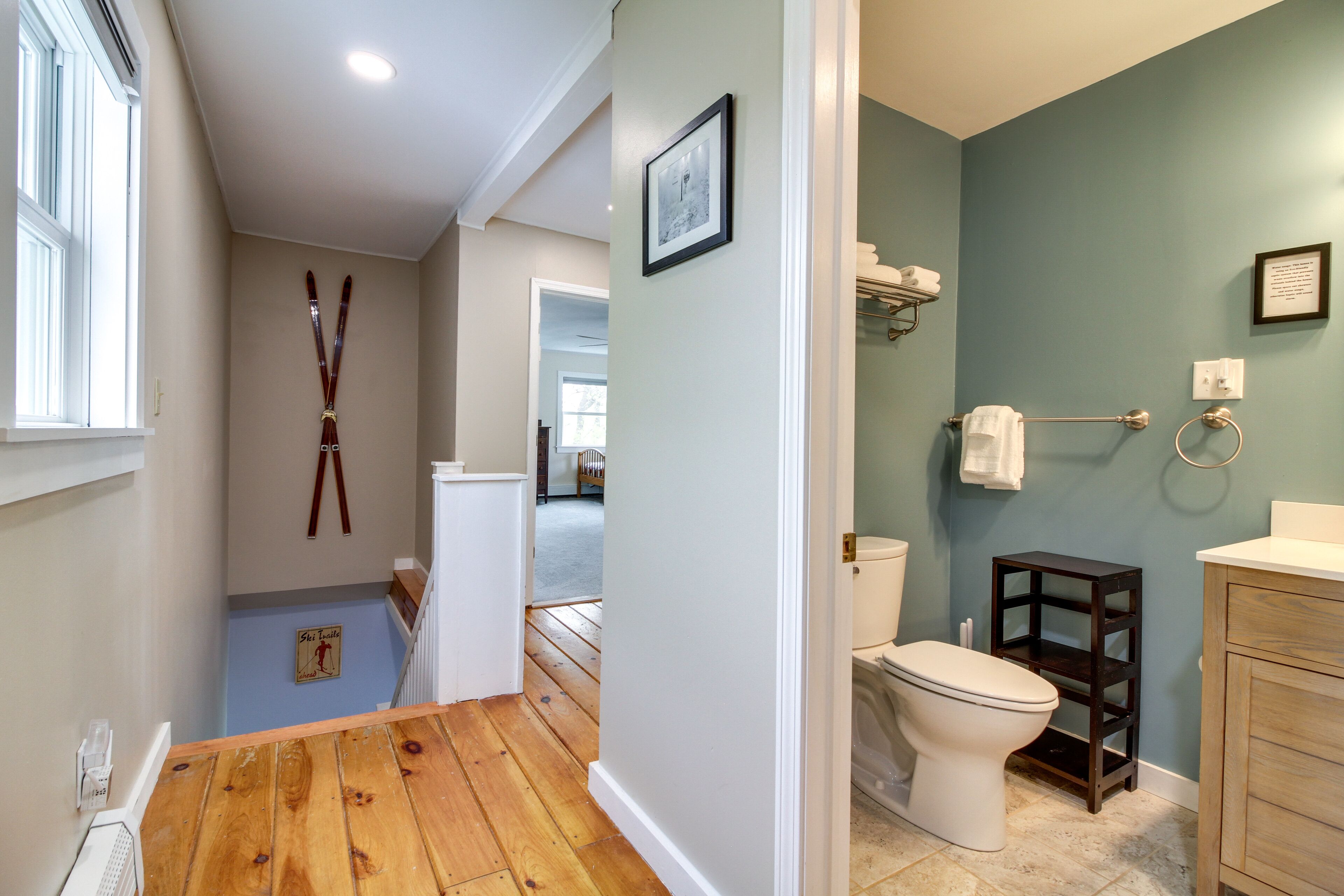 A bathroom with a toilet, sink, and a small shelf. There is a window with blinds and a picture on the wall.