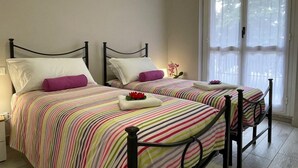A room with two beds, each with a black metal frame and striped bedding. There is a window with sheer curtains allowing natural light.