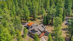 A large, two-story house surrounded by dense forest, with a view of snow-capped mountains in the background.