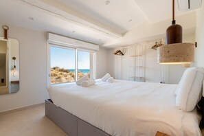 A bedroom with a large bed, a window with a view of the beach, a hanging light fixture, and a wardrobe.