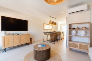 A modern living room with a flat-screen TV, a wooden cabinet, a round coffee table, and a kitchen area with bar stools.