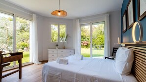 A bedroom with a large bed, a desk, and a view of a garden through the windows.