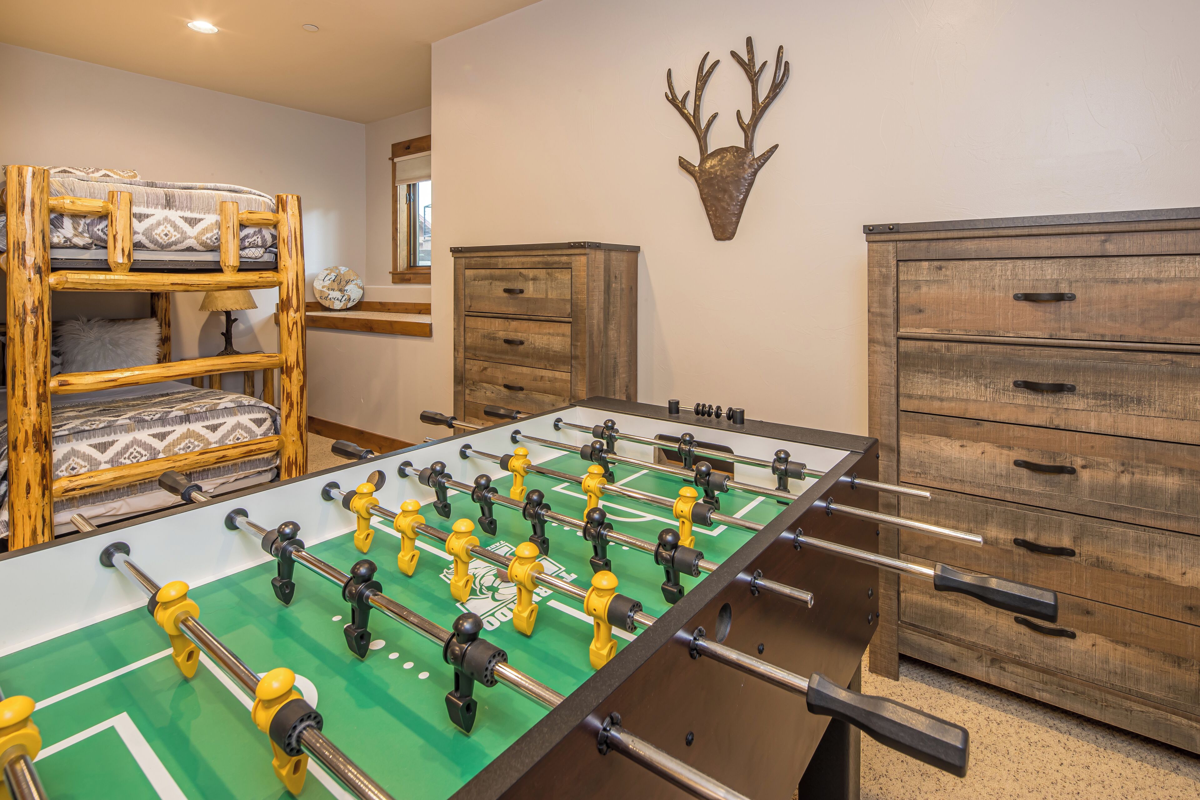 Una habitación con mesa de futbolín, literas, armarios de madera y un adorno de cabeza de venado en la pared.
