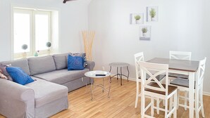 A living room with a grey sofa, a small round table, and a wooden dining table with white chairs. There are potted plants on the windowsill and a wall-mounted picture frame.