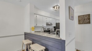 Una cocina moderna con armarios blancos, encimera de granito y campana extractora de acero inoxidable. Hay dos taburetes blancos de bar y un adorno mural.