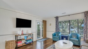 Una sala de estar con una televisión de pantalla plana, dos sillones azules, una mesa de centro blanca y un estante de madera con juegos de mesa.