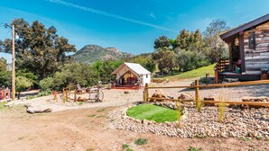 A rustic cabin with a porch, surrounded by a gravel area and a small lawn, set against a backdrop of hills and trees.