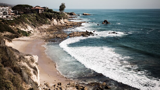 A coastal landscape with a sandy beach, rocky shoreline, and a few buildings on a cliff.