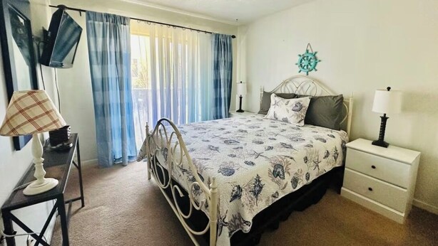 Quarto com cama, mesas de cabeceira, escrivaninha, cadeira, janela com cortinas e um adorno na parede.