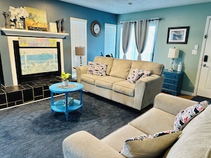 Una sala de estar con un sofá color crema, una mesa de centro azul, una chimenea y un televisor que muestra una escena de playa.