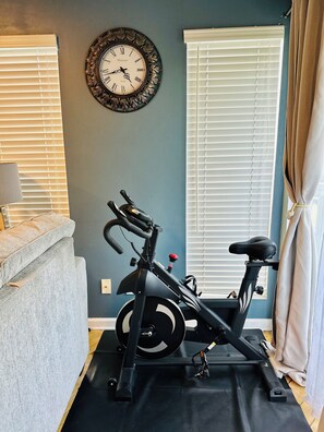 Una habitación con una bicicleta estática, un reloj en la pared y una ventana con persianas.