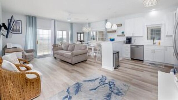 Una sala moderna con sofá, una mesa de centro y una silla de mimbre. Hay una zona de cocina con armarios blancos y una mesa de comedor.