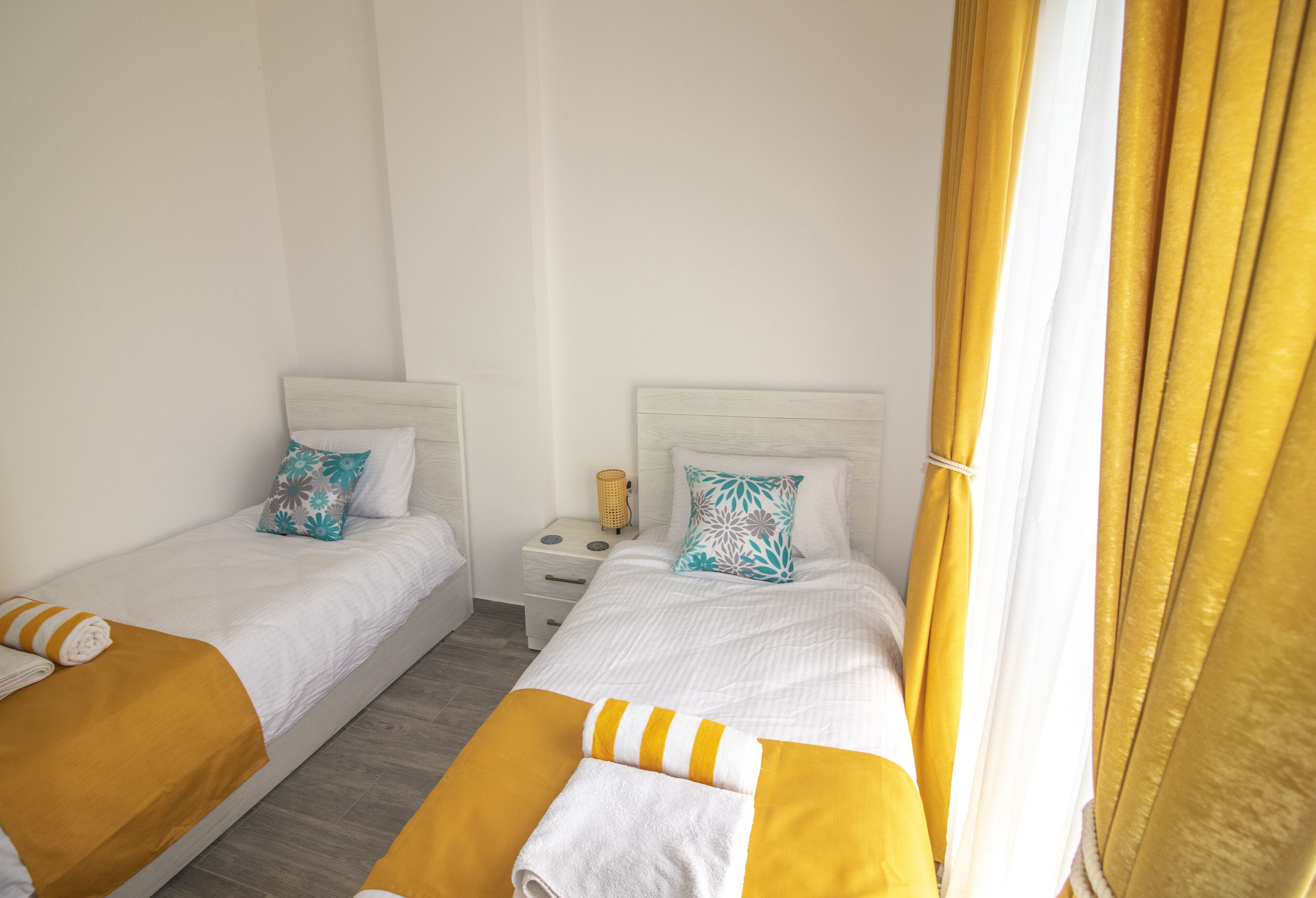 A hotel room with two single beds, each with a white headboard and a yellow accent pillow. There is a small bedside table with a lamp and a decorative item.
