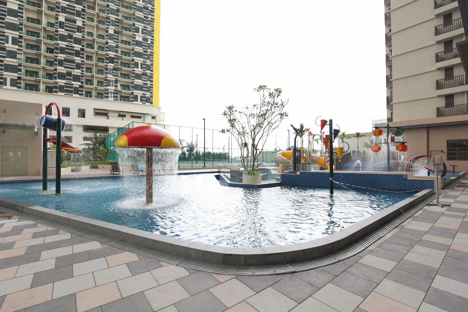 An outdoor swimming pool with water features, surrounded by a tiled walkway and adjacent buildings.