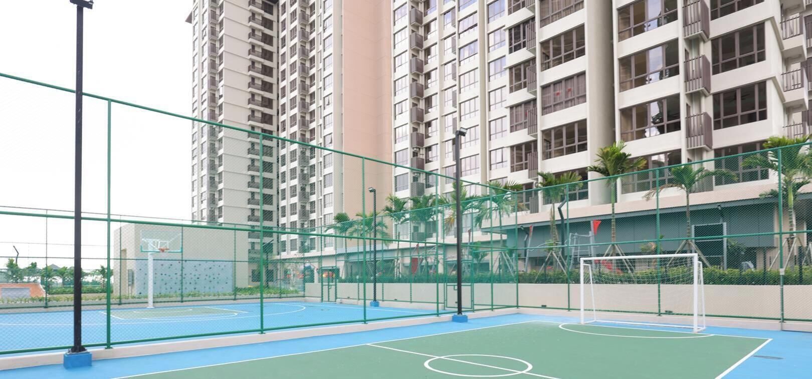 An outdoor basketball court with a basketball hoop and a soccer goal, surrounded by a green fence and tall residential buildings in the background.
