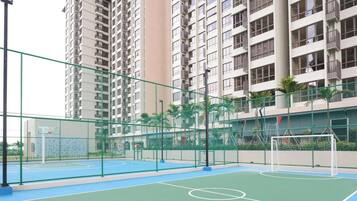 An outdoor basketball court with a basketball hoop and a soccer goal, surrounded by a green fence and tall residential buildings in the background.
