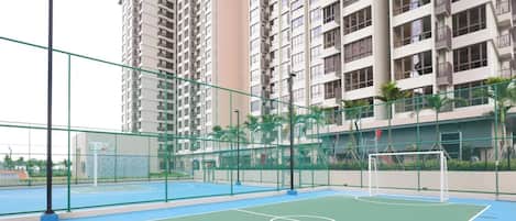 Uma quadra de basquete ao ar livre com um aro de basquete e um gol de futebol, cercada por uma cerca verde e edifícios residenciais altos ao fundo.