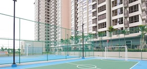 Una cancha de baloncesto al aire libre con un aro de baloncesto y una portería de fútbol, rodeada por una cerca verde y altos edificios residenciales al fondo.