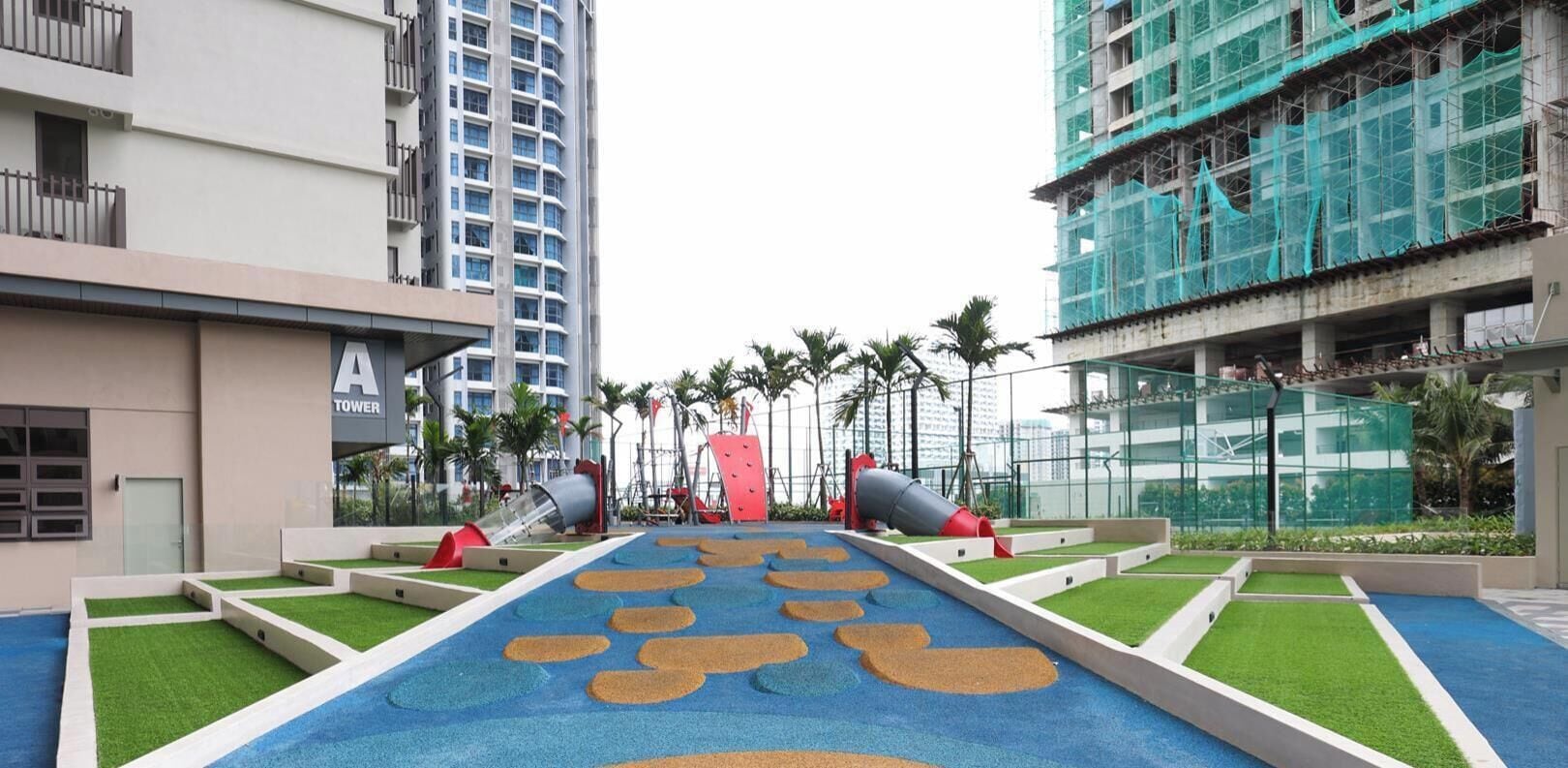 A playground area with slides and artificial grass, surrounded by modern buildings.