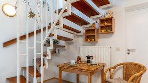 Una escalera con escalones de madera y barandillas blancas, una mesa de madera con tazas y una cafetera, un sillón de mimbre y una estantería con frutas.