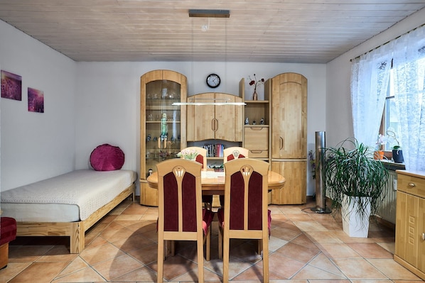 A compact living space with a bed, dining table, chairs, wooden cabinets, a clock, and a potted plant.