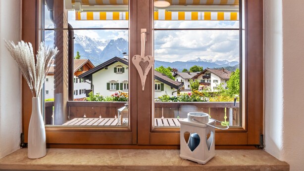 Una ventana con vista a casas y montañas, una decoración con forma de corazón y un jarrón con plantas secas.