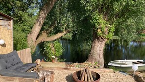 A wooden deck with a fire pit, a lounge chair, and a small boat by a lake. - Apartment by the Park Lake with Lake Access (Bad Bodenteich)