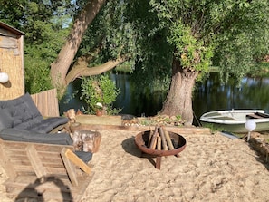 Una terraza de madera con un brasero, una silla de descanso y un pequeño bote junto a un lago.
