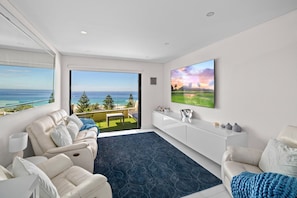 Un salon moderne doté d’une grande fenêtre donnant sur une plage, d’un téléviseur à écran plat et d’un meuble de rangement blanc pour les appareils multimédias.