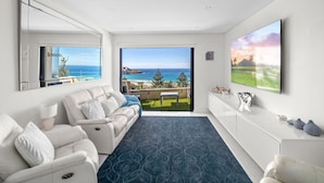 Ein modernes Wohnzimmer mit einem weißen Sofa, einem blauen Teppich mit Muster, einem großen Fernseher und einem Balkon mit Meerblick.