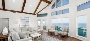 A spacious living room with a large window overlooking the ocean, a cozy seating area, and a wooden ceiling.