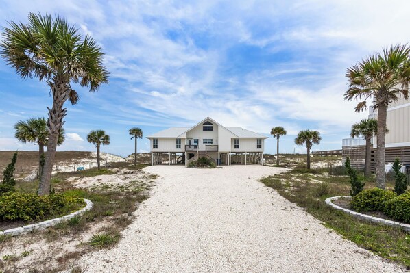 A two-story house with a gravel driveway, surrounded by palm trees and a sandy landscape.