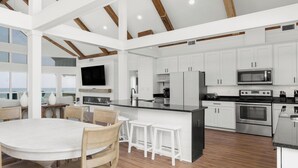A spacious kitchen with white cabinetry, a large island, and a dining area with a round table and wooden chairs. - Modern Beach Front Home with Private Beach Access | Trillion Dollar View by Kaiser (Gulf Shores)