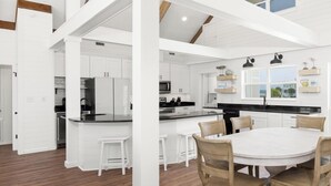 A spacious kitchen with white cabinetry, a central island, and a dining area with a round table and wooden chairs. - Modern Beach Front Home with Private Beach Access | Trillion Dollar View by Kaiser (Gulf Shores)