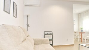Un salon avec un canapé beige, un climatiseur, un radiateur mobile et un coin repas comprenant une table et des chaises.