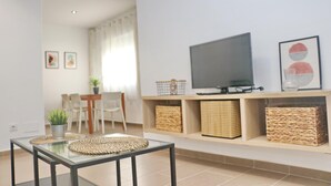 Un salon avec un téléviseur, des paniers en osier, une table basse en verre et, au fond, un coin repas.