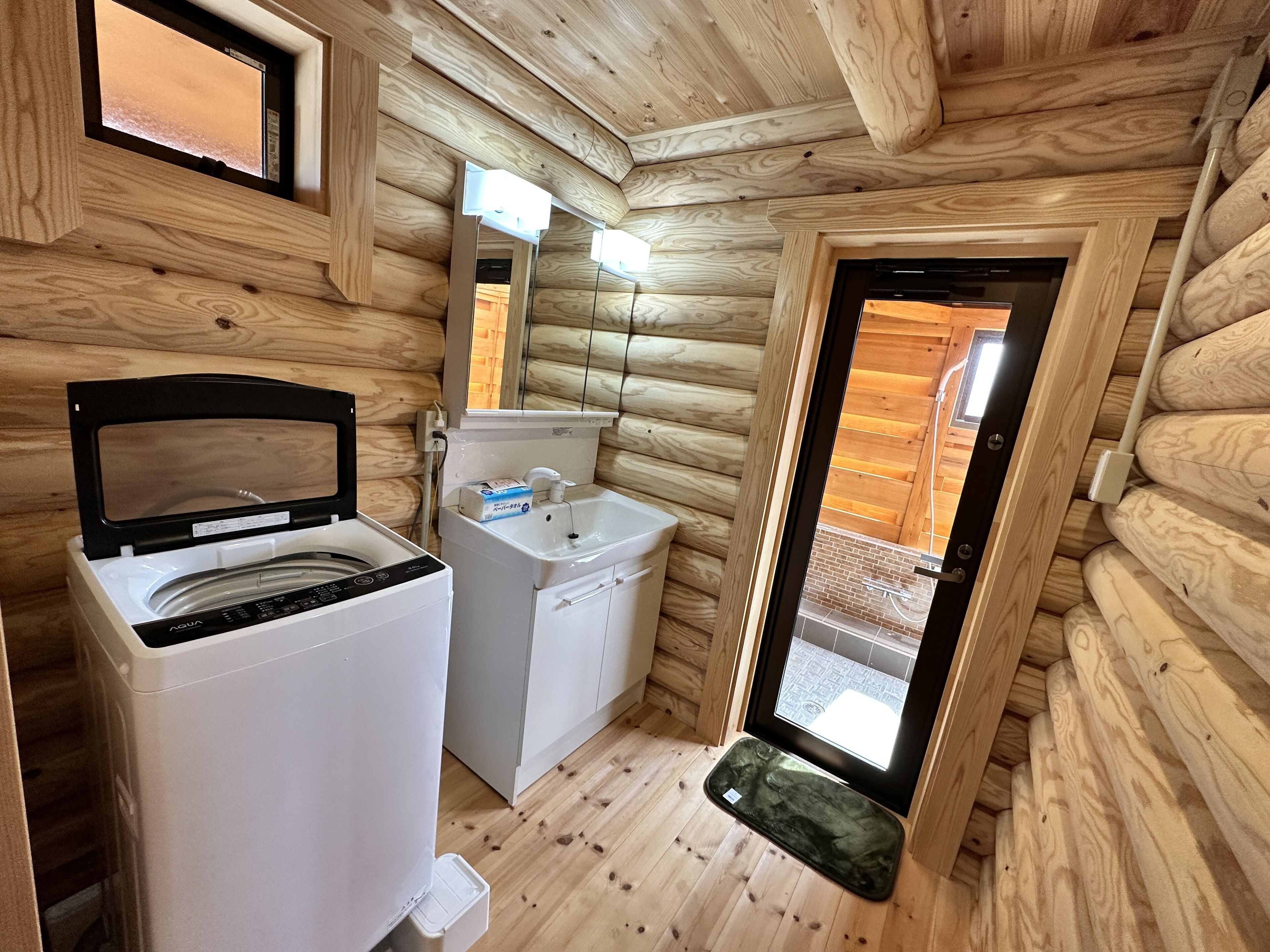 Een compact houten badkamer in een vakantiehuis, met een wasmachine, wastafel en een klein raam.