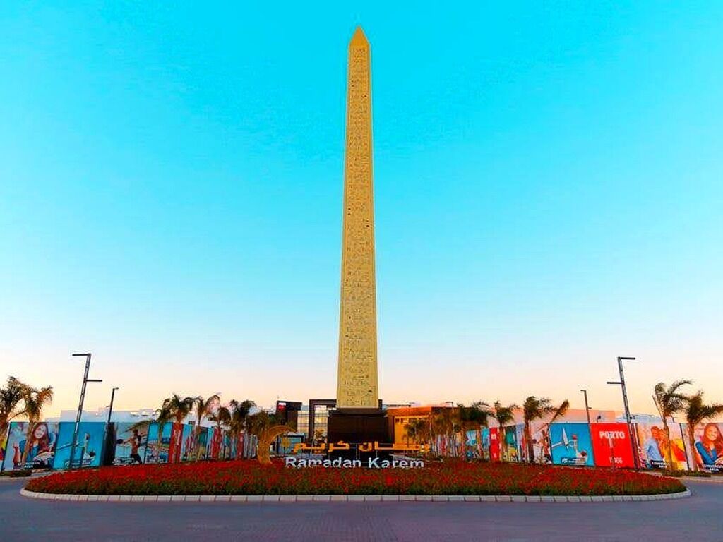 A tall obelisk monument with inscriptions, surrounded by a landscaped area with palm trees and a clear sky.