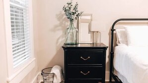 A bedroom with a black nightstand, a mirror, a vase with flowers, a basket with a towel, and a bed with white bedding. - 115 yr old Historic Heffner House in Downtown Rogers (Rogers)