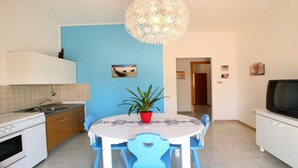 Une cuisine de taille réduite avec un mur bleu, une table à manger avec des chaises bleues, une plante en pot et un téléviseur posé sur un meuble.