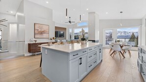A modern kitchen with a large island, white cabinetry, and a dining area with a view of mountains. - Luxury Kelowna House w/Pool and Sauna (West Kelowna)