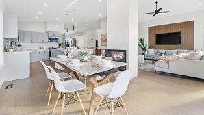 A modern kitchen with white cabinetry, a dining area with a white table and chairs, and a living room with a sofa and TV. - Luxury Kelowna House w/Pool and Sauna (West Kelowna)