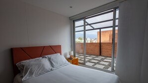 A bedroom with a bed, a nightstand, and a large window with a view of a brick wall and buildings. - Apartamento con Terraza Privada en la Calleja. A 3 min de la Clinica Reina Sofía (Bogotá)