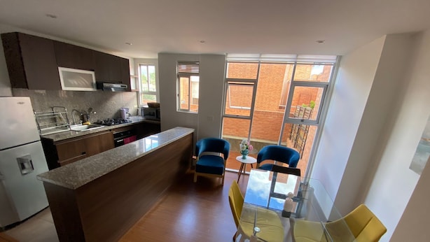 A modern kitchen with a central island, a dining area with a glass table and chairs, and large windows offering a view of the outside. - Apartamento con Terraza Privada en la Calleja. A 3 min de la Clinica Reina Sofía (Bogotá)