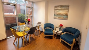 A dining area with a glass table, yellow and black chairs, and a blue armchair. - Apartamento con Terraza Privada en la Calleja. A 3 min de la Clinica Reina Sofía (Bogotá)