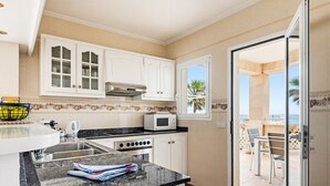 Une cuisine avec des meubles blancs, un four à micro-ondes et une cuisinière. Il y a une fenêtre donnant sur des palmiers et un balcon avec des chaises.